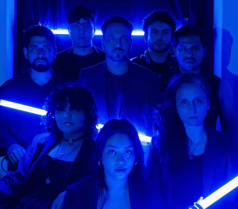 Ocho personas en formación, con vestimenta oscura y luces azules, creando un ambiente artístico y misterioso.