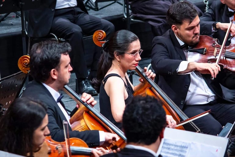Músicos en chaquetas negras y camisas blancas tocan en un escenario oscuro; la solista de chelo destaca en el centro.