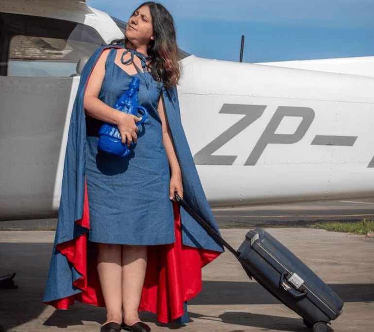 Mujer en vestido azul y capa roja, sosteniendo un objeto azul, se muestra decidida junto a un avión pequeño en un entorno despejado.