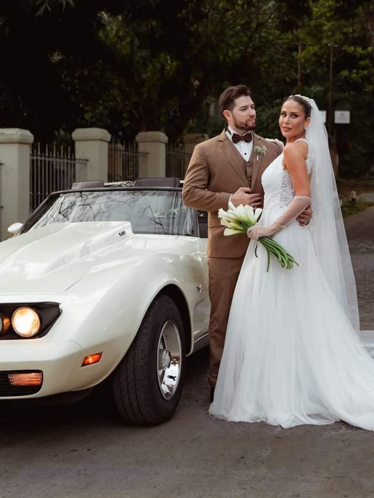 Hombre de traje marrón y mujer de vestido de novia blanco sonríen junto a un automóvil deportivo en una vía pública.