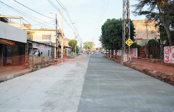Calle en construcción con asfalto nuevo, acera sin terminar, árboles y señales de tráfico visibles.
