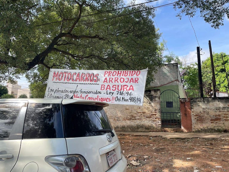 Cartel en cerca de ladrillos que dice 'MOTOCARROS PROHIBIDO ARROJAR BASURA' en ambiente soleado y urbano.