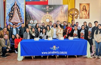 Grupo de personas atentas en evento formal, con decoraciones religiosas y documentos en la mesa.