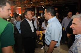 Miguel Prieto en traje oscuro y Ricardo Estigarribia en camiseta verde se saludan en un ambiente festivo con fondo de asistentes.