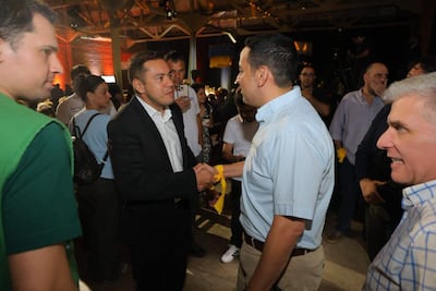 Miguel Prieto en traje oscuro y Ricardo Estigarribia en camiseta verde se saludan en un ambiente festivo con fondo de asistentes.