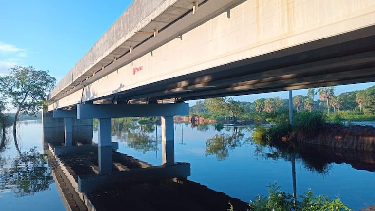Puente de concreto elevado sobre el agua, rodeado de vegetación, con un reflejo nítido y sin personas visibles en la escena.