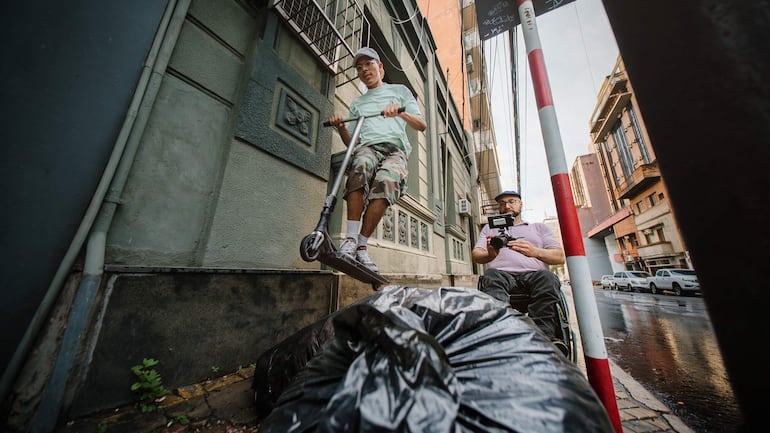 Un joven en camiseta verde claro y pantalones cortos camuflados patina mientras un hombre en silla de ruedas sostiene una cámara.