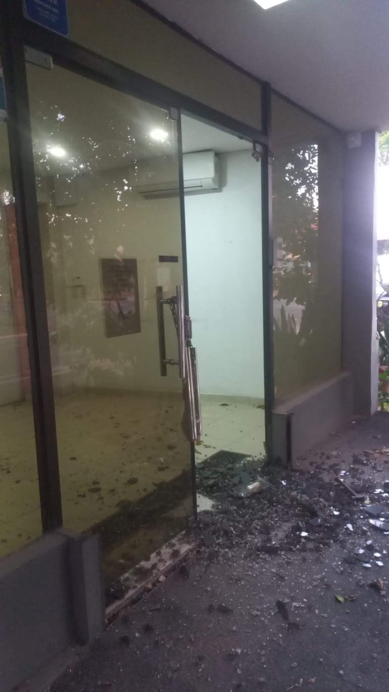 Puerta de vidrio parcialmente abierta con cristales rotos en el suelo, indicando un posible acto de vandalismo en un entorno urbano.