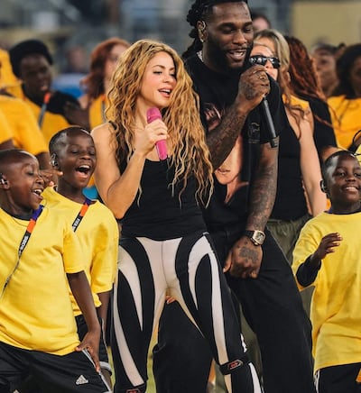 Shakira con cabello ondulado y micrófono rosa, cantando junto a Burna Boy y niños con camisetas amarillas, en un ambiente festivo.
