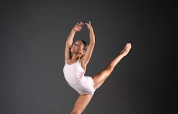 Bailarina en leotardo blanco salta elegantemente en un fondo negro, cuerpo en movimiento y brazos extendidos.
