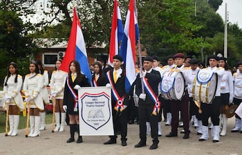 Estudiantes en uniformes formales desfilan, sosteniendo una bandera del Colegio Nacional 'Puerto Guaraní'.