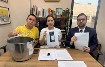 Johanna Ortega en blusa blanca sostiene un documento, mientras Bruno Martinez en camiseta amarilla y Maximiliano Seiferheld en traje oscuro observan atentos.
