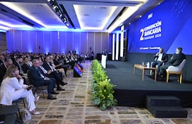Benigno López gesticula en el escenario mientras Dimas Ayala escucha atentamente. Auditorio moderno lleno de público formal.