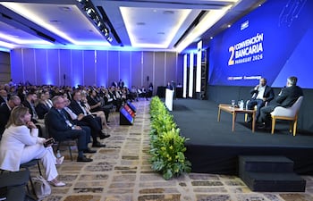 Benigno López gesticula en el escenario mientras Dimas Ayala escucha atentamente. Auditorio moderno lleno de público formal.