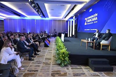 Benigno López gesticula en el escenario mientras Dimas Ayala escucha atentamente. Auditorio moderno lleno de público formal.