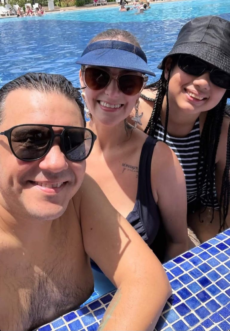 Hombre sin camiseta con gafas de sol sonríe junto a dos mujeres, una en traje de baño y otra en camiseta a rayas, junto a una piscina.