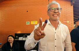 Rigoberto Chamorro, con camisa blanca y sonrisa, levanta dos dedos mientras se encuentra en una cantina con paredes de ladrillo naranja.