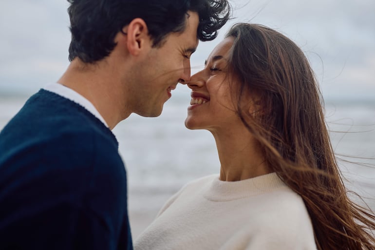 Hombre y mujer sonríen en la playa, el hombre con suéter oscuro y líneas claras, la mujer con suéter blanco, muy cerca uno del otro.