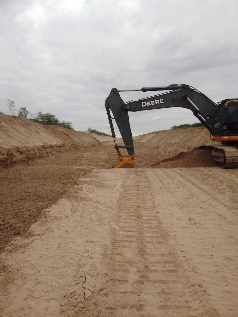 Excavador Deere trabajando en terreno arenoso sin presencia de personas, con cielo nublado y vegetación escasa al fondo.