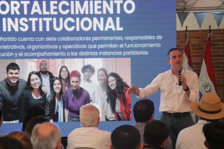 Stephan Rasmussen con camisa blanca y micrófono, mientras el público lo escucha atentamente en un salón decorado con banderas.