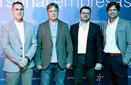 Hombre de pie, camisa blanca y saco gris, sostiene micrófono mientras habla ante una multitud en evento corporativo.