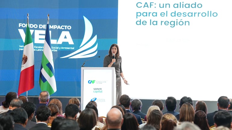 Mujer oradora en un auditorio con micrófono, audiencia atenta y banderas de México y logo de CAF al fondo.