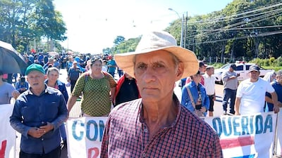 Hombre mayor con sombrero habla en manifestación, mientras una mujer de blusa colorida sostiene un cartel en un ambiente cívico.