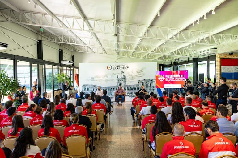 Audiencia en camisetas rojas aplaudiendo mientras un hombre habla con micrófono en un evento formal.