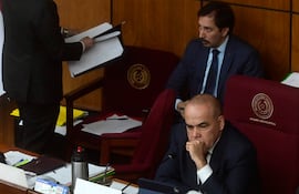 Basilio Gustavo Núñez Giménez en traje oscuro, con expresión pensativa, rodeado de otros hombres formales en ambiente legislativo.