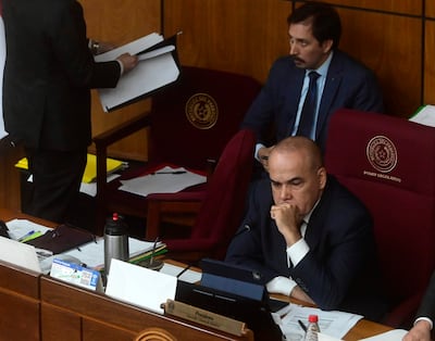 Basilio Gustavo Núñez Giménez en traje oscuro, con expresión pensativa, rodeado de otros hombres formales en ambiente legislativo.