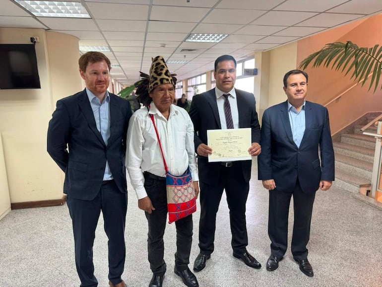 Cuatro hombres en un entorno formal; uno en traje oscuro sostiene un certificado mientras otro lleva indumentaria tradicional con sombrero de plumas.