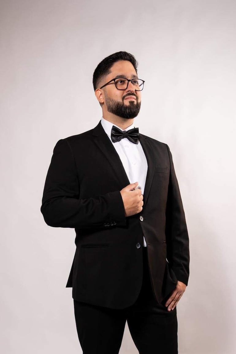 Hombre de pie con traje negro y pajarita, mirada reflexiva y barba cuidada en fondo claro.
