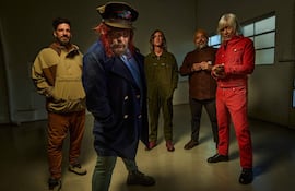 Cinco hombres posan en estudio; uno con abrigo azul y gorro, otro en overol verde, y otro en camisa marrón.