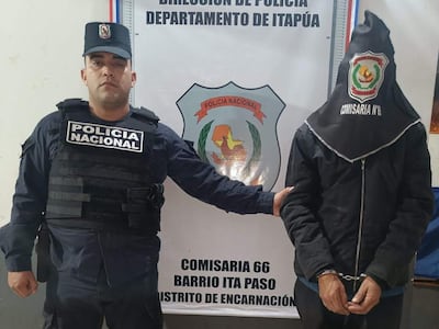 Oficial de policía en uniforme sostiene a un hombre con capucha, cara visible incluso esposado, en la Comisaría 66 de Ita Paso.