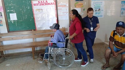 Hombre mayor en silla de ruedas, con camisa de cuadros, es ayudado por mujer en camiseta roja en sala blanca.