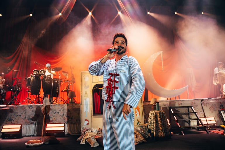 Cantante en pijama a rayas azules y blancas sosteniendo un micrófono, rodeado de músicos en un escenario con cortinas rojas.
