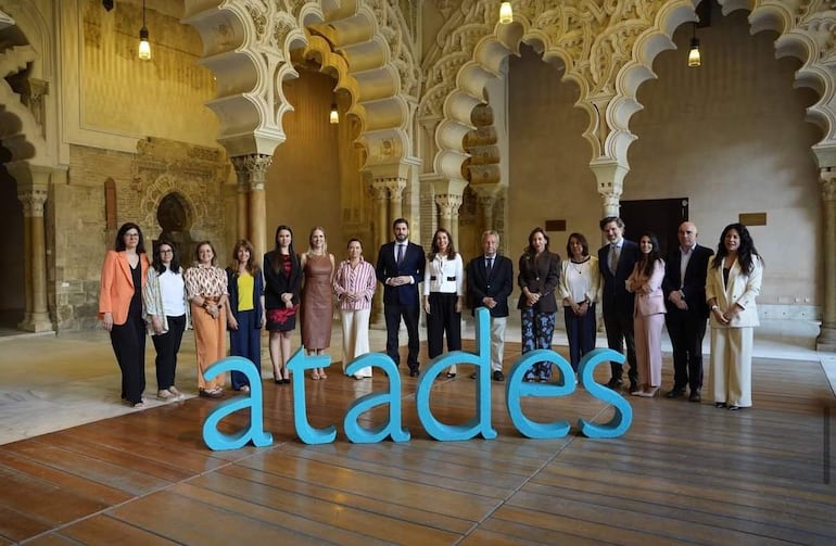 Grupo de 13 personas, la mayoría mujeres con vestidos coloridos y un hombre en traje oscuro, sonriendo en un entorno arquitectónico notable.
