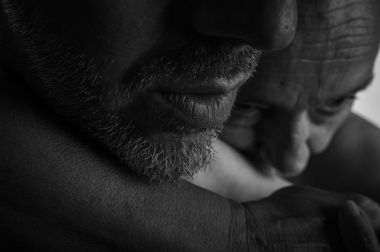 Hombre con barba canosa abraza al otro con cercanía, en un ambiente de estudio fotográfico con fondo oscuro.