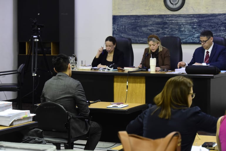 Hombre de espaldas con traje oscuro, tres autoridades judiciales revisando documentos y tomando notas en un entorno sobrio.