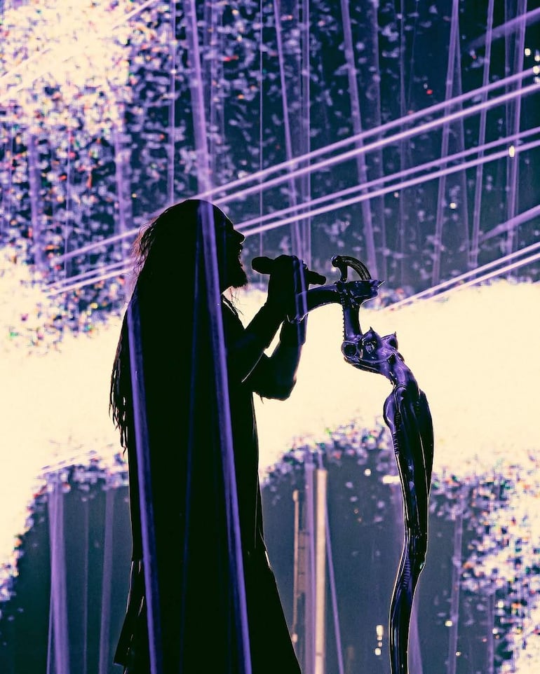 Jonathan Davis de Korn en el escenario, con cabello largo y suelto, sosteniendo un micrófono frente a un ambiente de luces brillantes.