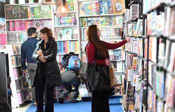 Mujer con abrigo negro sostiene un libro, otra mujer en blusa roja revisa estantería, hombre de espaldas observa libros.