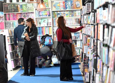 Mujer con abrigo negro sostiene un libro, otra mujer en blusa roja revisa estantería, hombre de espaldas observa libros.
