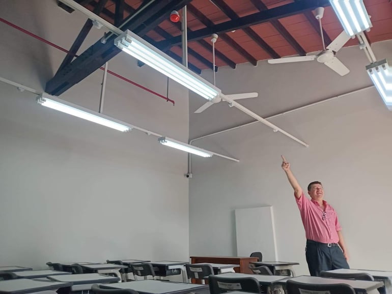 Hombre con camisa rosa y pantalones oscuros, de pie en aula moderna, señalando el techo, rodeado de escritorios y sillas.