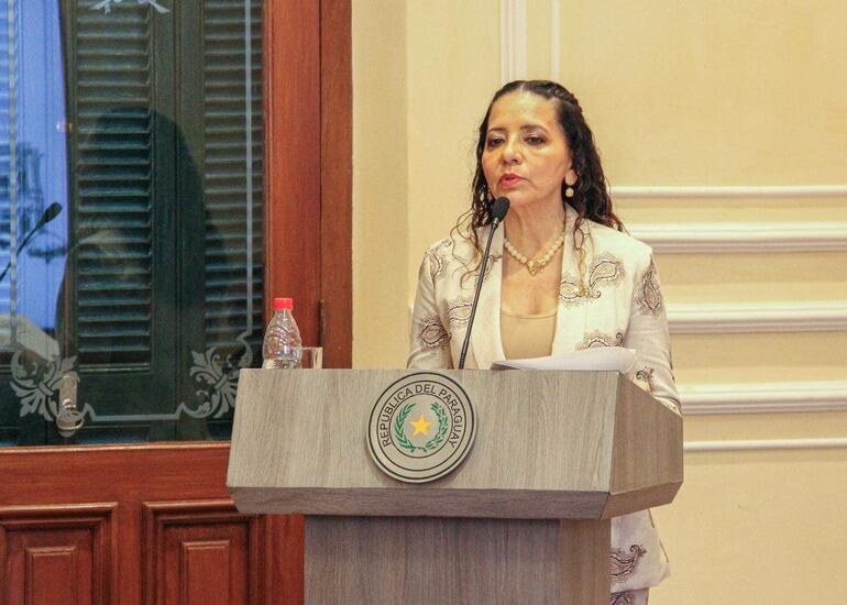 Lourdes González en chaqueta clara y collar de perlas habla en podio con el escudo de Paraguay mientras sostiene documentos.