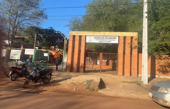Persona vestida de oscuro camina hacia la escuela con vehículos estacionados y árboles alrededor.
