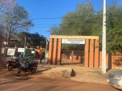 Persona vestida de oscuro camina hacia la escuela con vehículos estacionados y árboles alrededor.