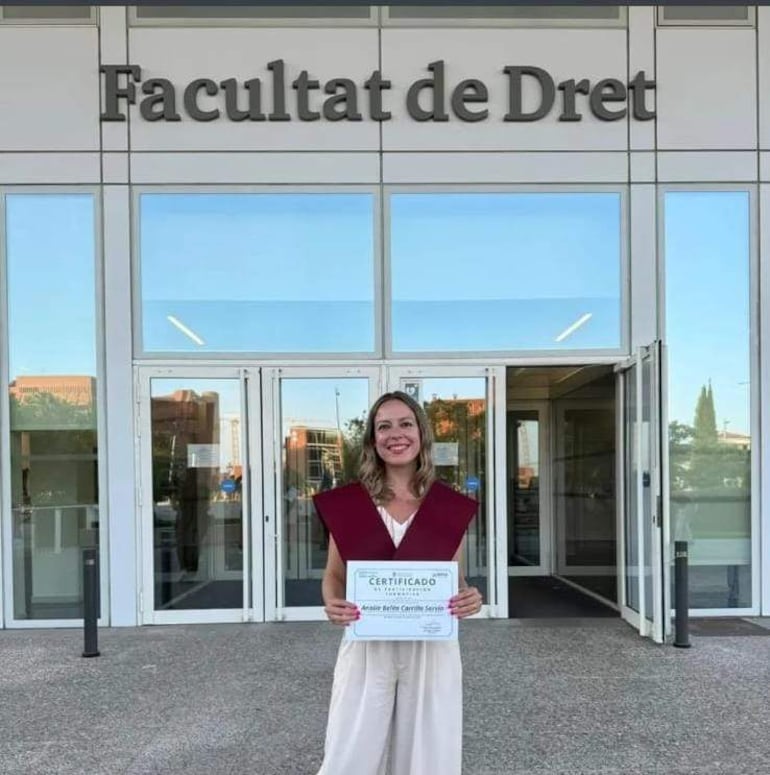 Mujer de pie con vestimenta académica burdeos, sonriente, sosteniendo un certificado frente a un edificio de la facultad.