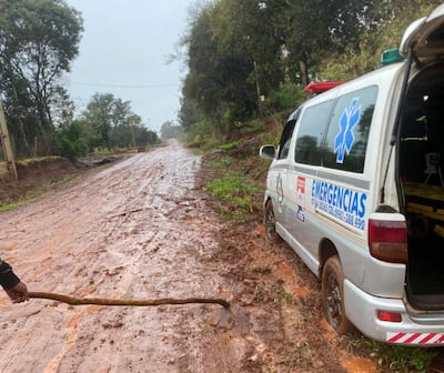 Ambulancia blanca con símbolo de emergencias atascada en barro, mientras un hombre sostiene un palo evaluando la situación.