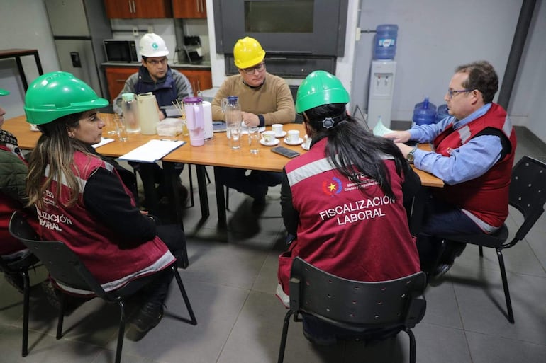 Cinco personas con chalecos de 'FISCALIZACIÓN LABORAL' discuten alrededor de una mesa, con cascos de protección y documentos en la sala.