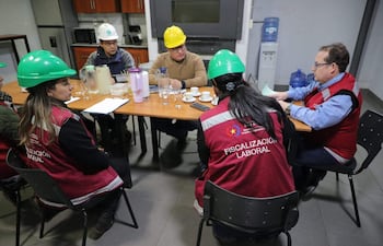 Cinco personas con chalecos de 'FISCALIZACIÓN LABORAL' discuten alrededor de una mesa, con cascos de protección y documentos en la sala.